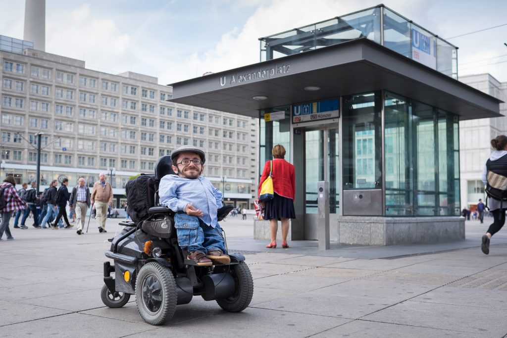 Rollstuhlfahrer Raul Krauthausen steht vor einem aufzug auf dem alexanderplatz in Berlin und guckt in die Kamera
