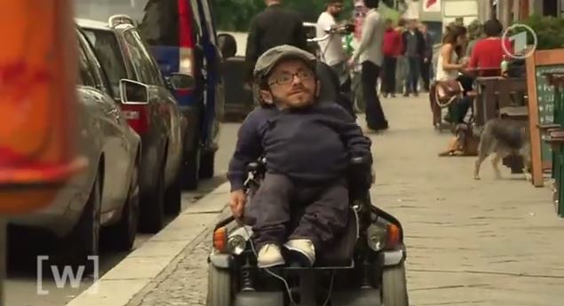 Standbild eines ARD Beitrages, auf dem Raul Krauthausen zu sehen ist, während er auf einem Gehweg mit dem Rollstuhl fährt