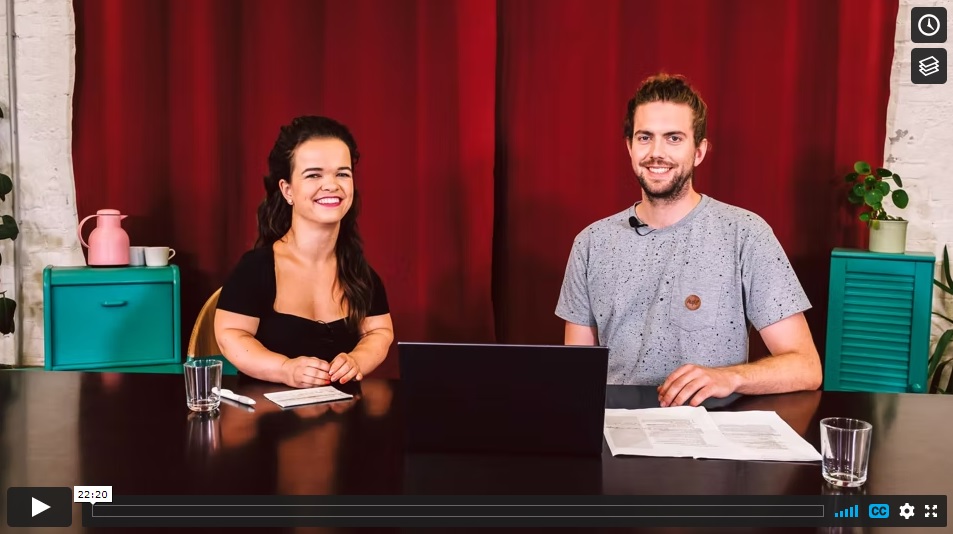 Screenshot eines Vimeo-Videos. Zwei Personen sitzen vor einem roten Samtvorhang an einem Tisch. Links eine kleinwüchsige Frau, rechts ein Mann. Beide lächeln in die Kamera. In der Mitte auf dem Tisch steht ein Laptop.