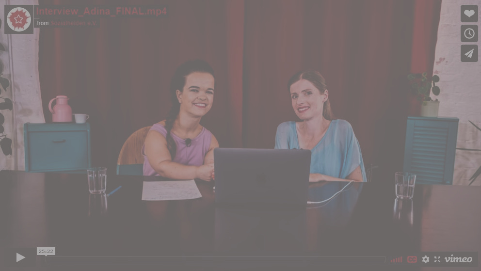 Zwei Frauen sitzen an einem Tisch vor einem roten Samtvorhang. Sie lächeln beide in die Kamera. In der Mitte auf dem Tisch steht ein Laptop.