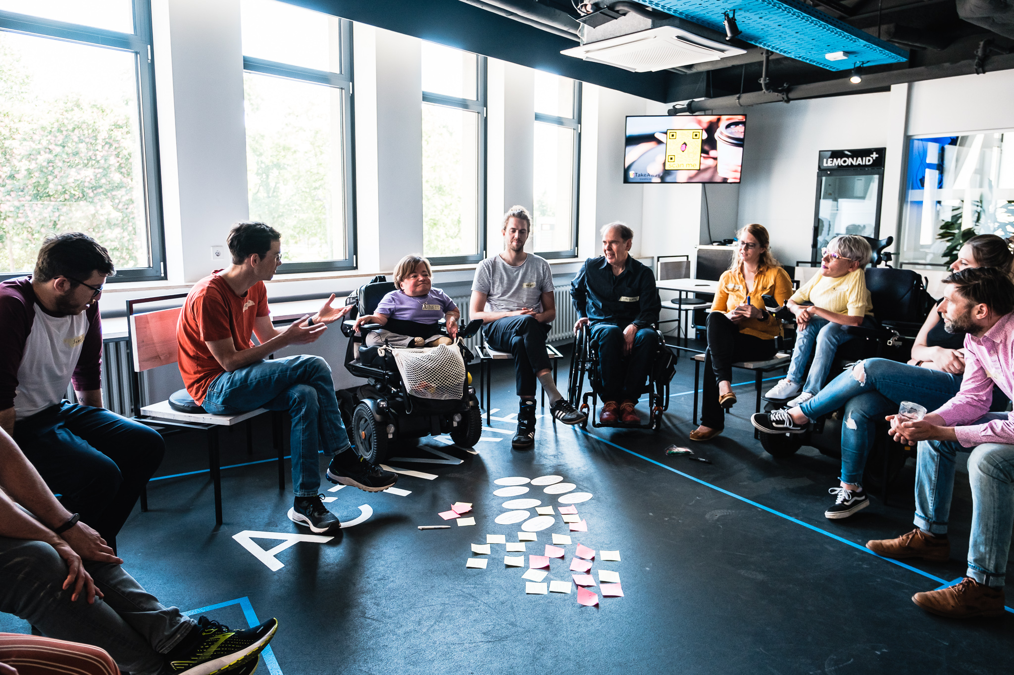 Foto von der Ideenwerkstatt in Leipzig, Die Teilnehmenden sitzen in einem Stuhlkreis. In der Mitte auf dem Boden liegen viele bunte Zettel.