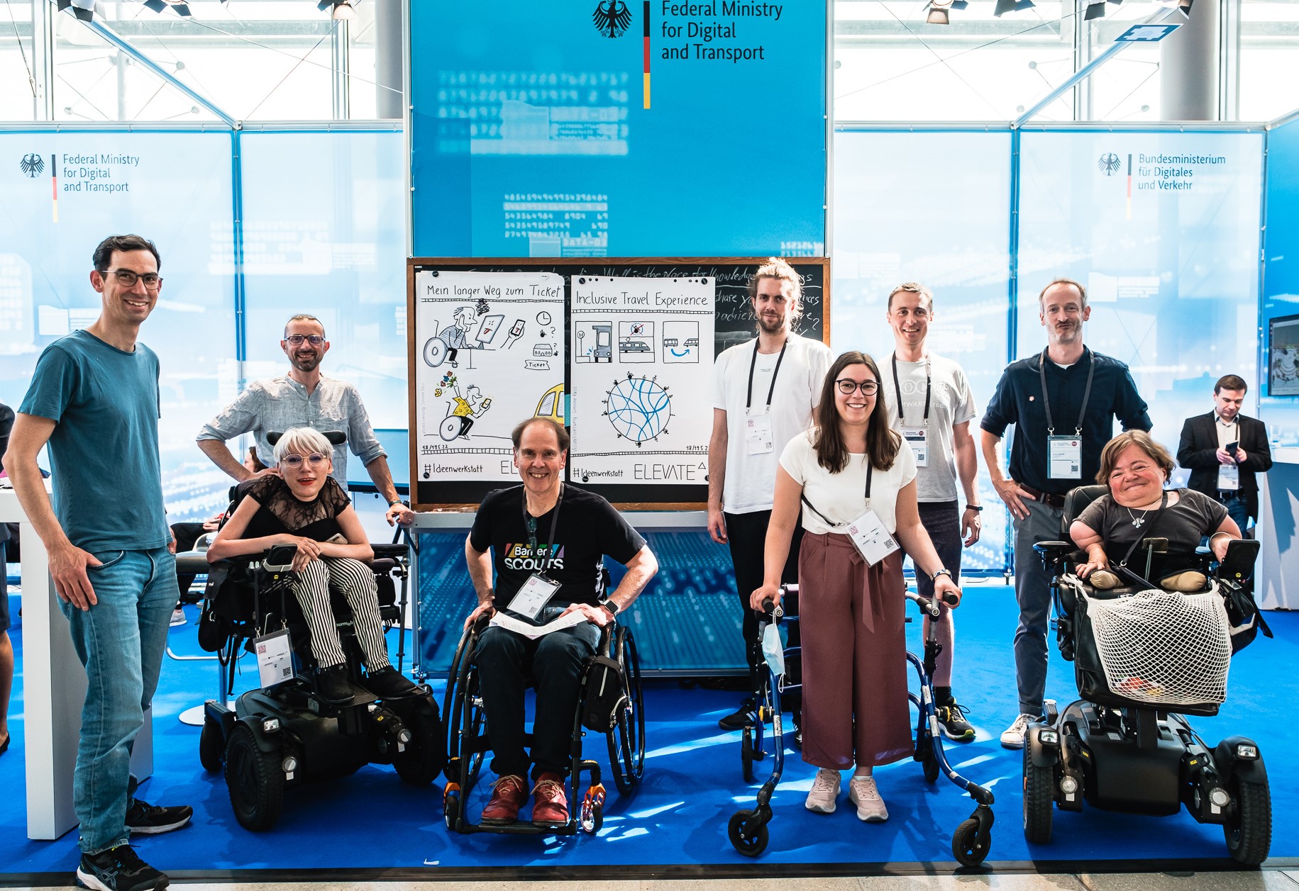 Eine Gruppe von Personen steht an einem Messestand auf der IFT 2022 in Leipzig. 3 Personen sitzen in einem Rollstuhl, eine Person steht an einem Rollator. Alle lächeln in die Kamera. Im Hintergrund ein Banner des Bundesministeriums für Digitales und Verkehr.