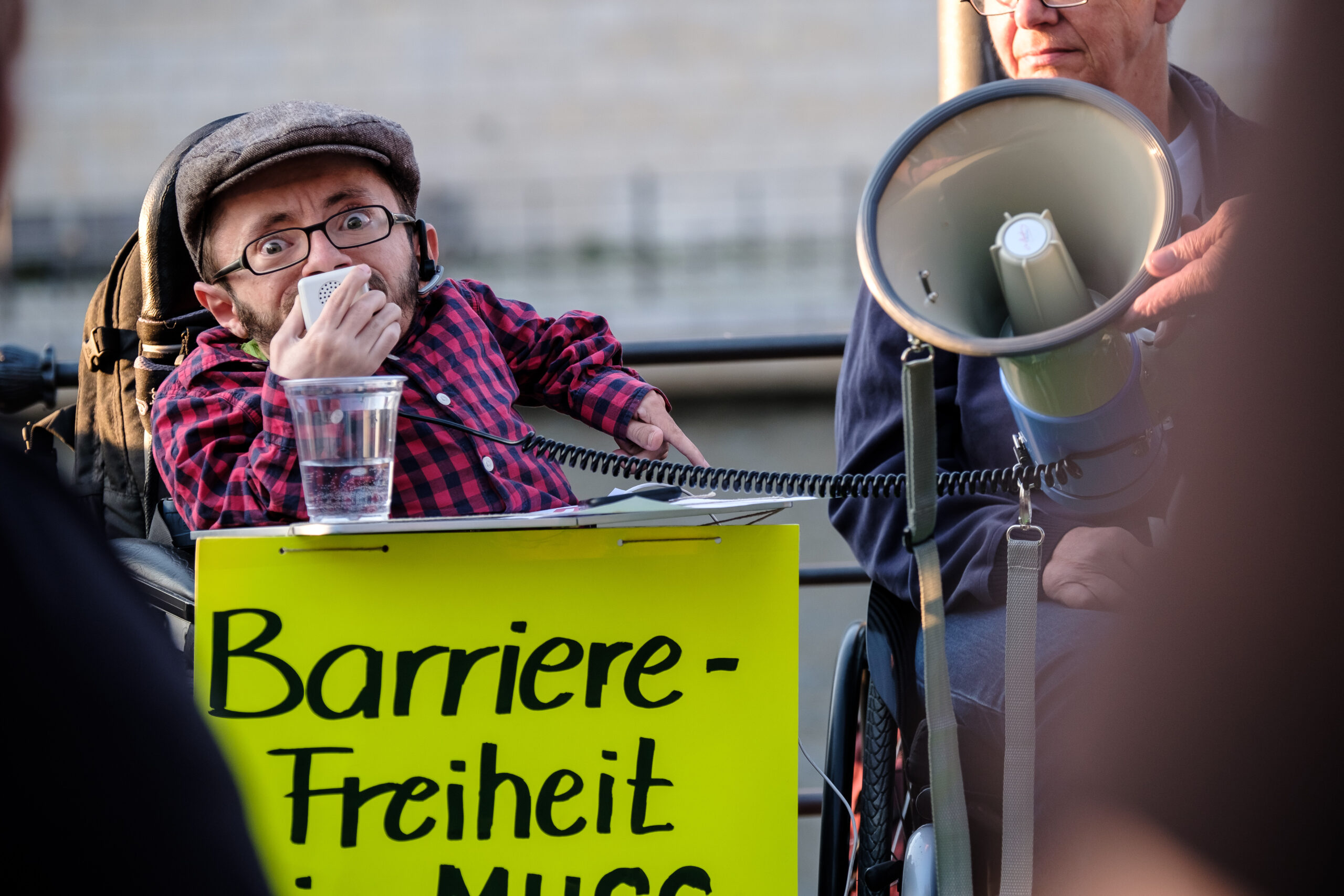 Foto von Raul Krauthausen. Er spricht in ein Megaphone. Am Tisch seines Rollstuhls ist ein Plakat angebracht auf dem steht "Barrierefreiheit".